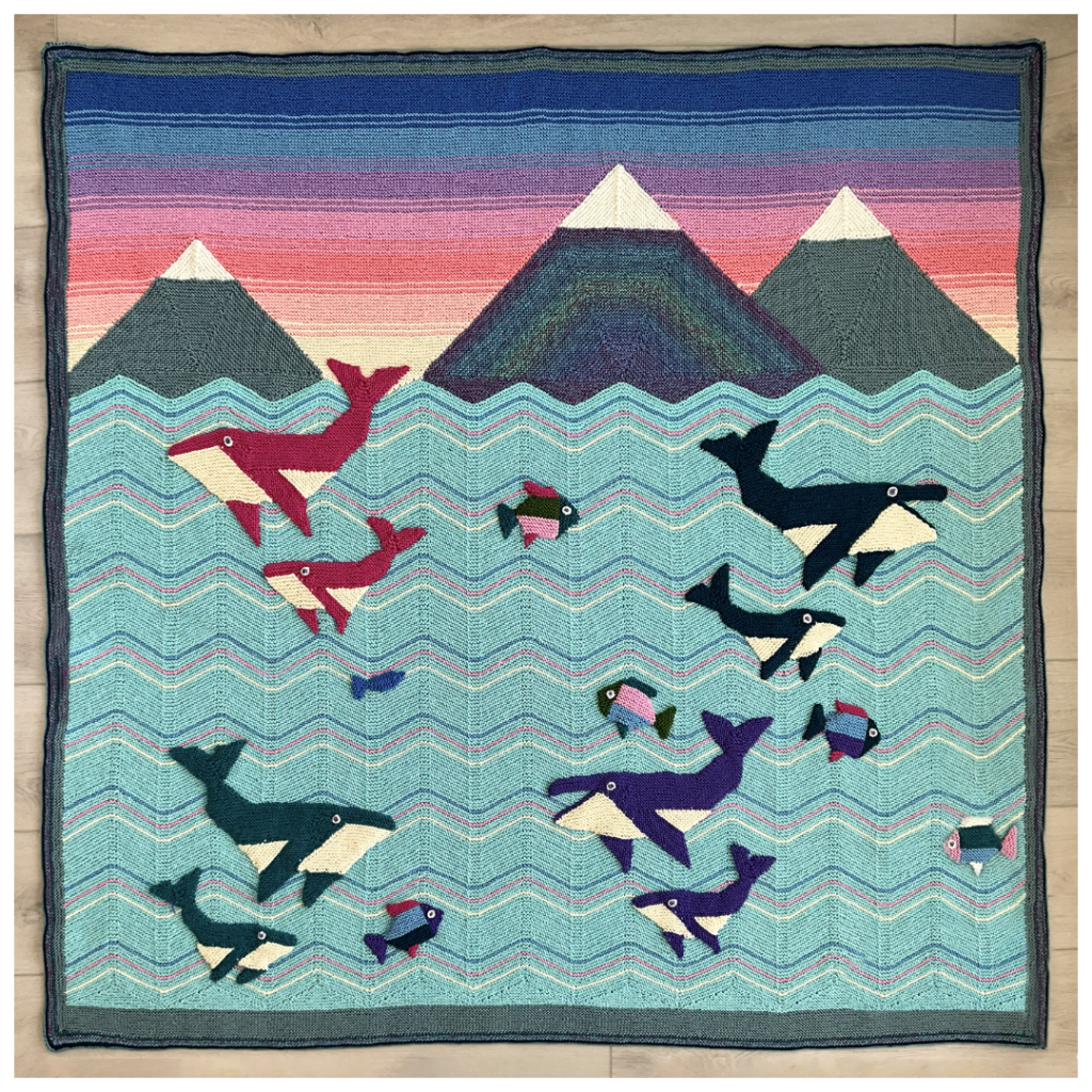 A knitted blanket featuring whales in various colors with waves and mountains. The Whales blanket is a design by Margaret Holzmann.