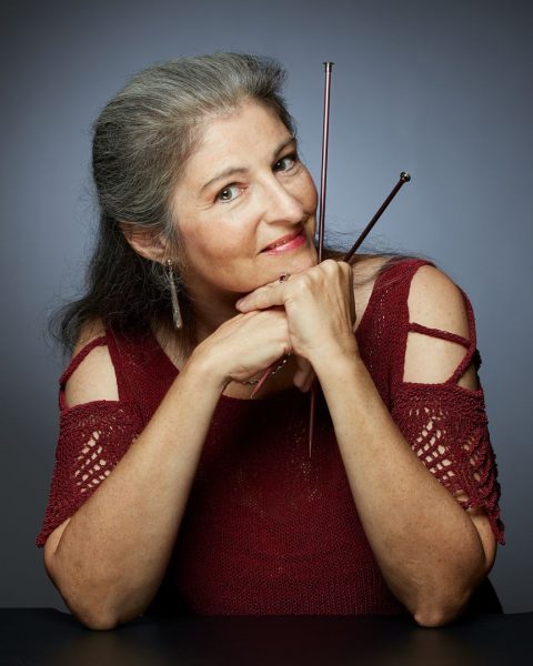A headshot of knit designer, Margaret Holzmann. She's wearing a red top, holding knitting needles, in front of a gray background.