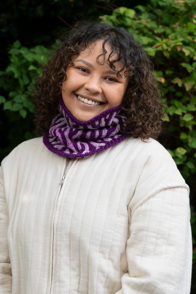 A person is standing outside wearing a white jacket with a light and dark purple cowl. The Up in Flames Cowl designed by Jennifer Kent is a free Cascade Yarns pattern.