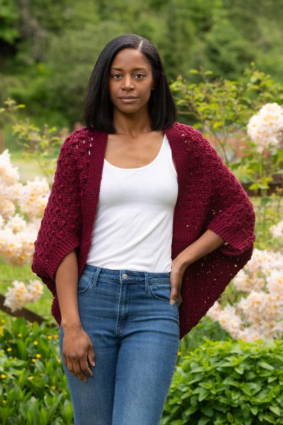 A person is standing outside wearing a red shrug. The Crochet Knit Shrug designed by Jennifer Kent is a free Cascade Yarns design.