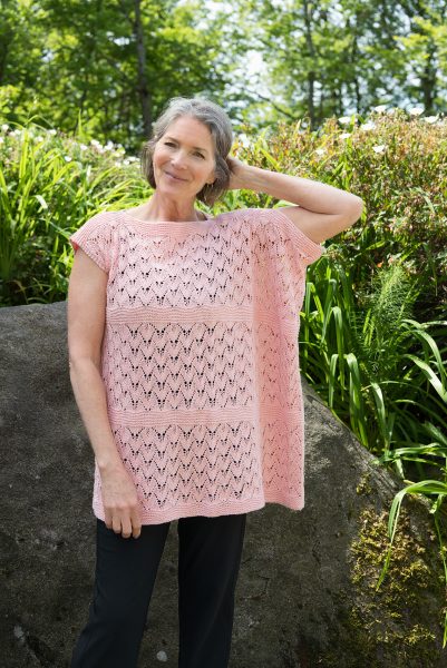 A person is standing outside wearing a light colored knit tee. The Perennial Tee designed by Jennifer Kent is a free Cascade Yarns.